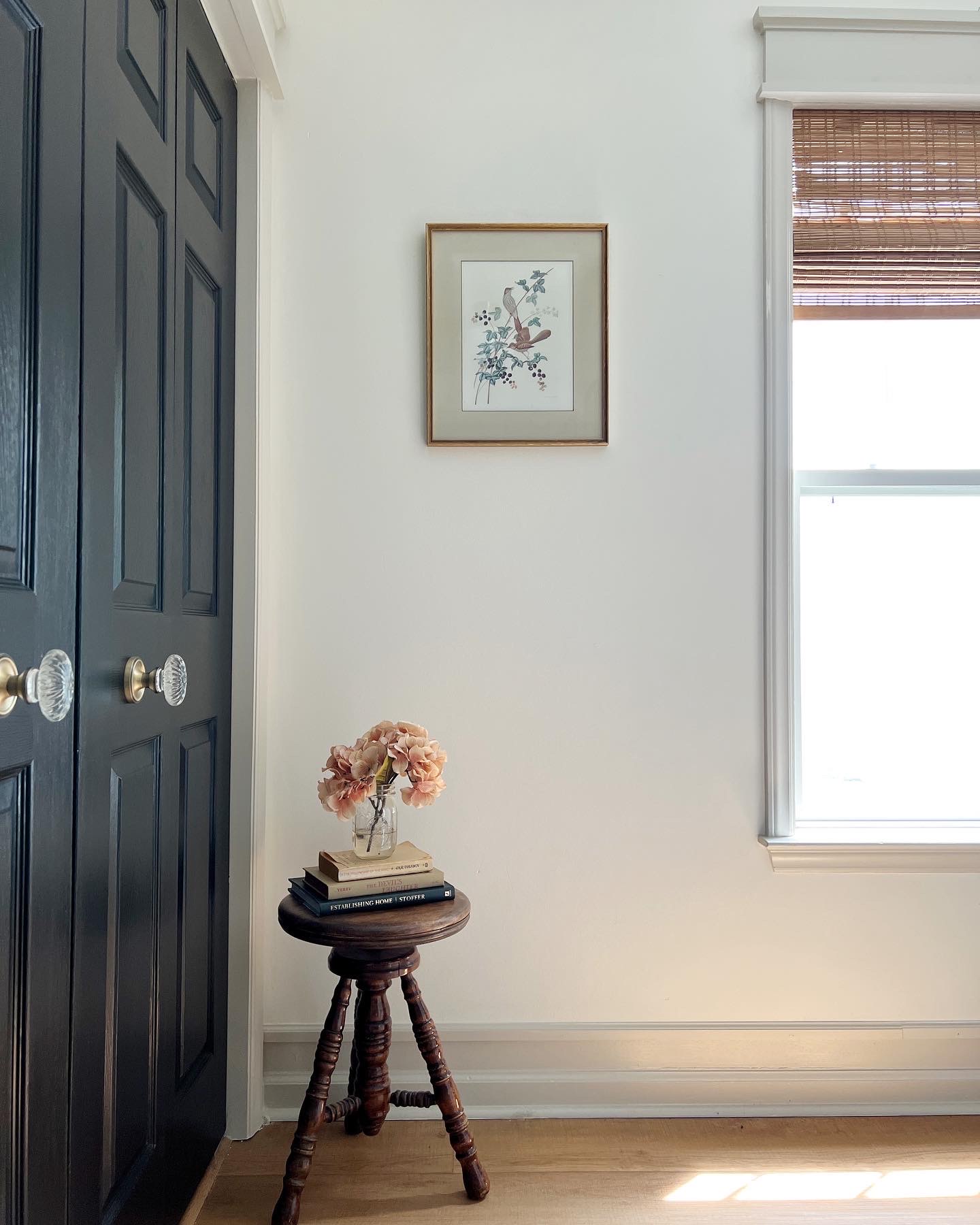 contrasting taupe trim in hallway. thrifted art hanging on th wall above wooden vintage piano stool with hydrangeas Black painted bi-fold doors with nostalgic warehouse knobs - gold rosette with crystal oval knobs. Craftsman style window trim covered in bamboo shade